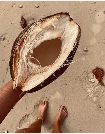 Et billede taget oppefra af en kvinde, der står med en halv kokosnød og hendes fødder i sandet er i baggrunden