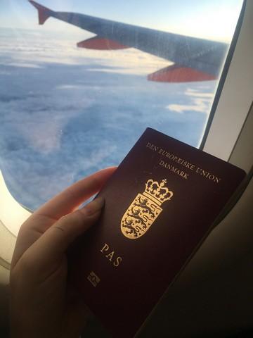 En kvindehånd holder hendes pas op mod ruden i flyet, hvor man i baggrunden kan se flyvingen over de flotte skyer