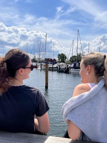  Zwei junge Mädchen, die durch Boblberg gute Freundinnen geworden sind, sitzen zusammen in der Sonne am Hafen und unterhalten sich.