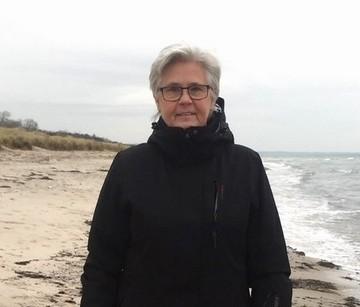  Ada, 65 år gammal, står och ler på stranden i en svart jacka med havet i bakgrunden och hon är glad eftersom hon har hittat nya väninnor på Boblberg efter en skilsmässa.