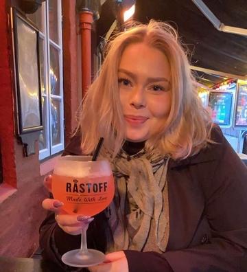 Line med lyst pagehår sidder på en bar med en drink i hånden og smiler, fordi hun har fundet nye veninde på Boblberg efter hun har været i et dårligt miljø