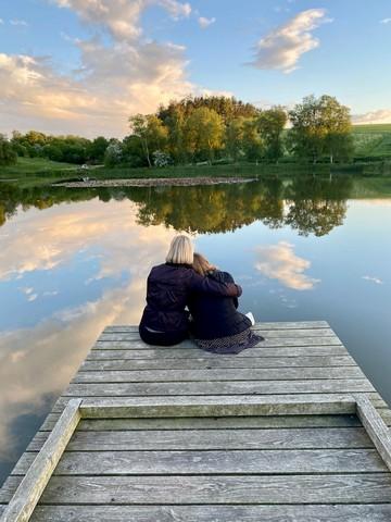 Zwei Frauen sitzen auf einem Badesteg, halten einander fest und schauen auf das Wasser und die Natur.