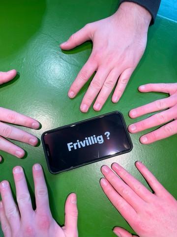 En telefon ligger på et bord og viser "frivillig" på skærmen, imens forskellige person har deres hånd omkring telefonen for at vise sammenhold og fællesskab