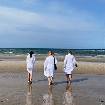 Drei Freundinnen, die sich auf Boblberg getroffen haben, gehen nebeneinander am Strand in weißen Morgenmänteln.