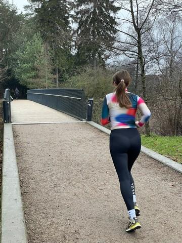 En mørkhåret ung kvinde i spraglet træningstøj er ude at løbe en tur i en park. hvor der er en bro henover søen