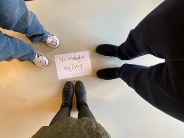Et billede taget oppefra af tre personer, som står omkring et hvidt papir, hvor der står "vi mangler dig"