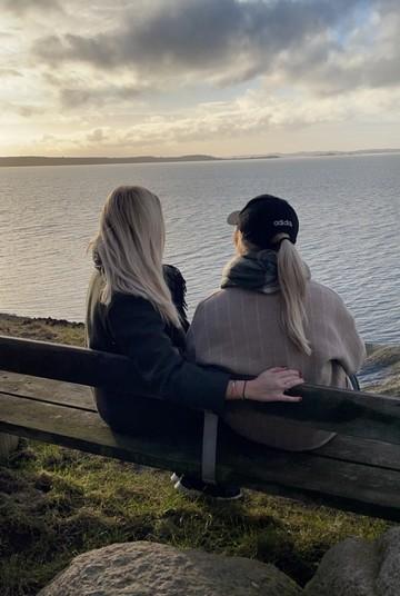 To piger sidder tæt på hinanden på en bænk ved havet og snakker om ensomhed